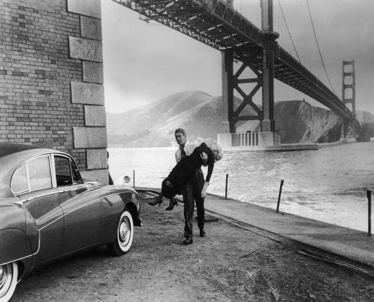 Beneath a soaring bridge, a man in shirtsleeves and tie carries a woman in heels and gloves. 