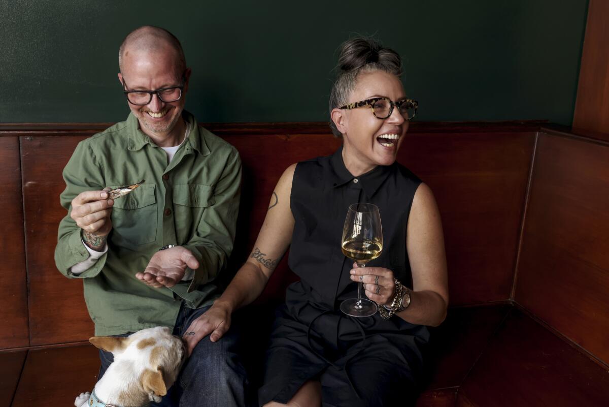 A laughing man holds a piece piece of fish and a laughing woman holds a glass of wine, while a dog looks on. 