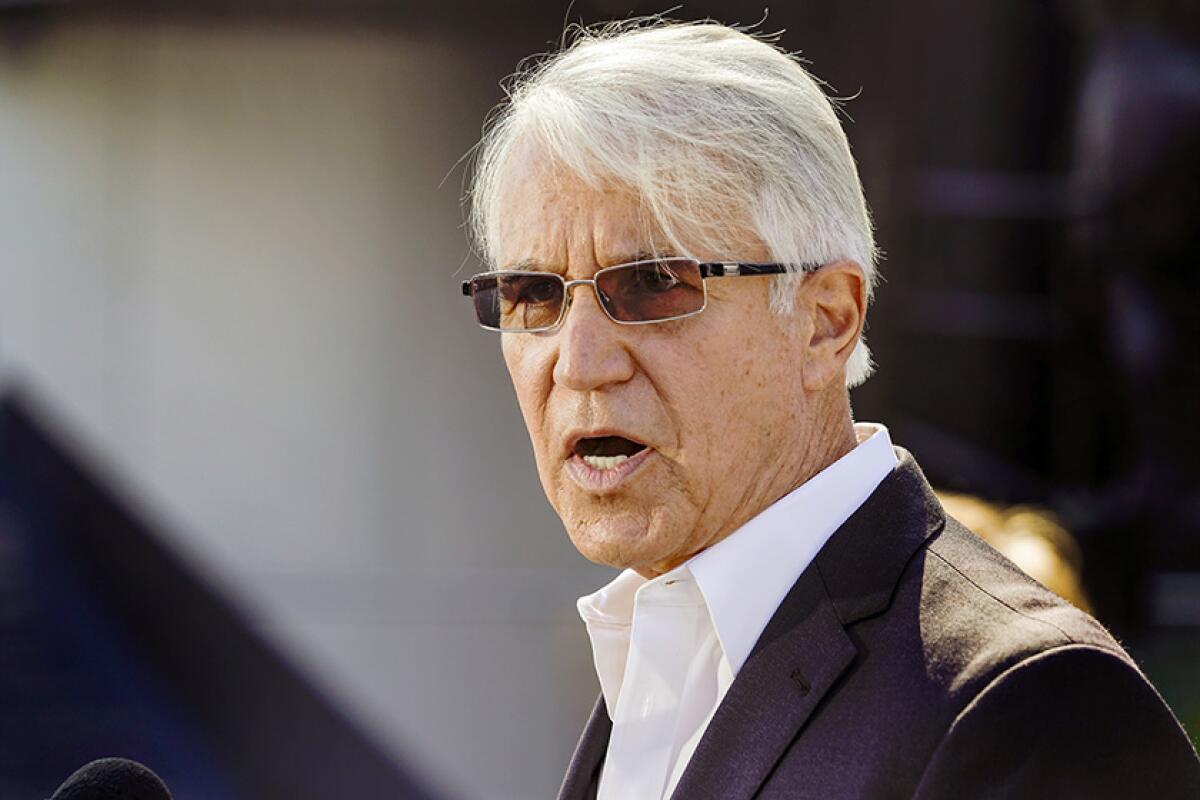 A close-up photo of a man with white hair, wearing sunglasses, a white shirt and a suit coat. 