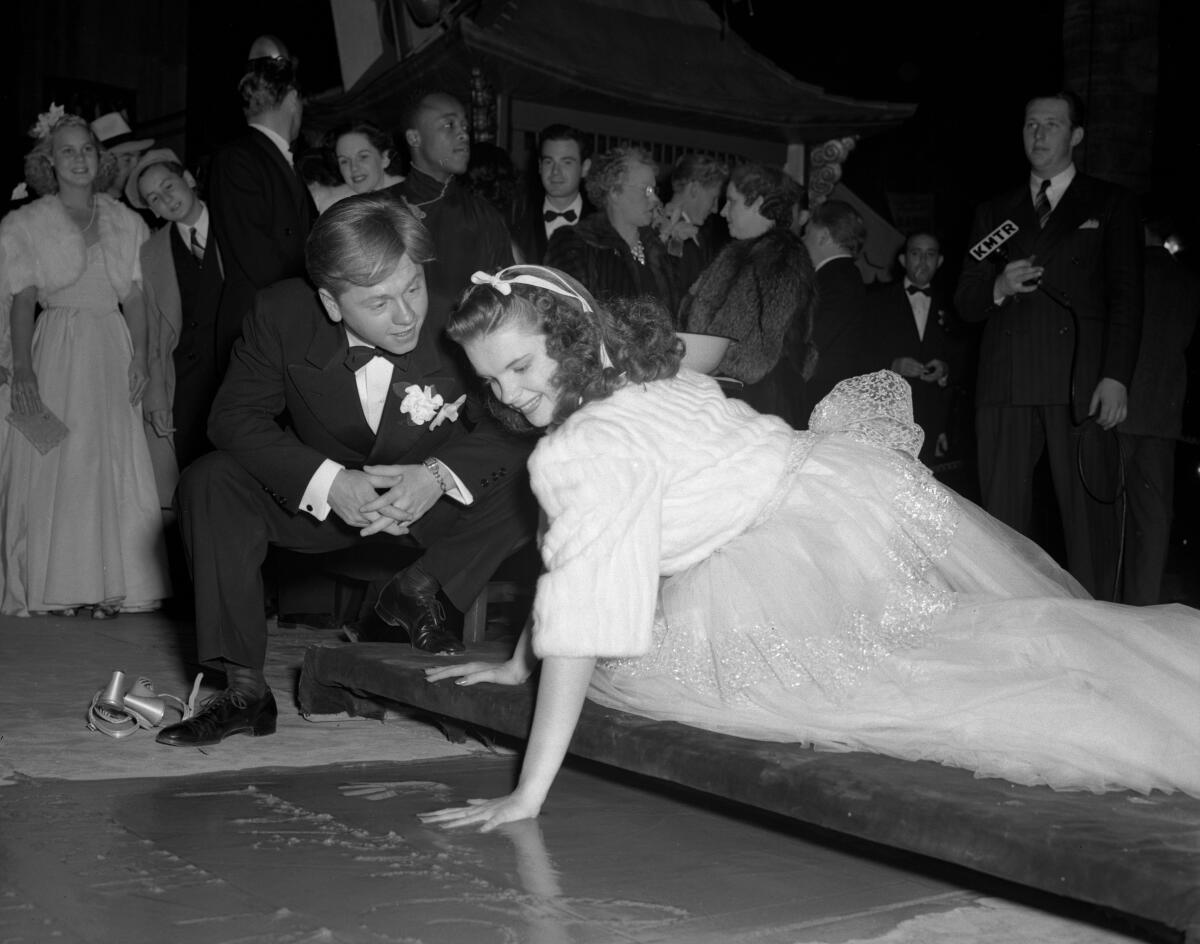 A young woman in tulle skirt and fur jacket presses her hand into wet cement. A man in formalwear crouches alongside her.