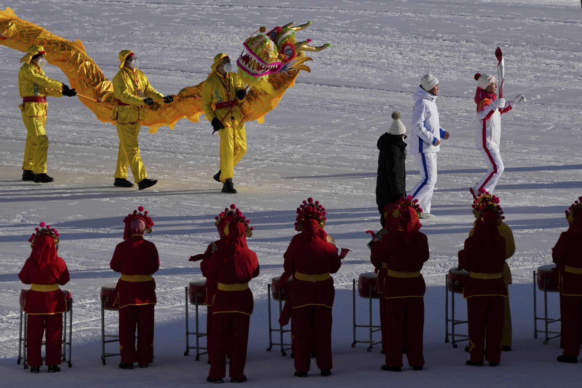 A person holds a torch next to another person. Behind, dancers in yellow costumes perform a dragon dance 