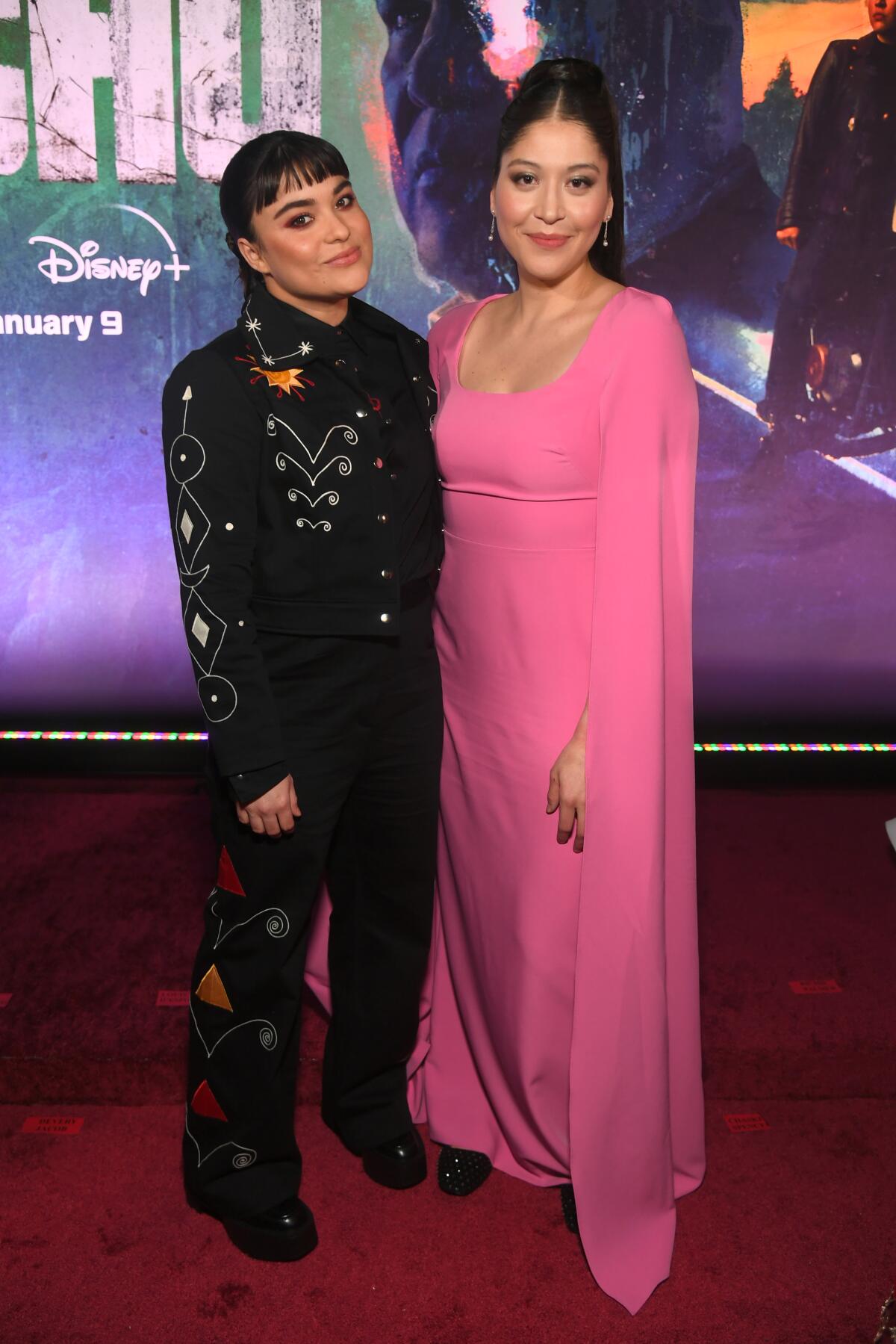Devery Jacobs stands in a black suit with abstract designs next to Alaqua Cox, clad in a pink gown