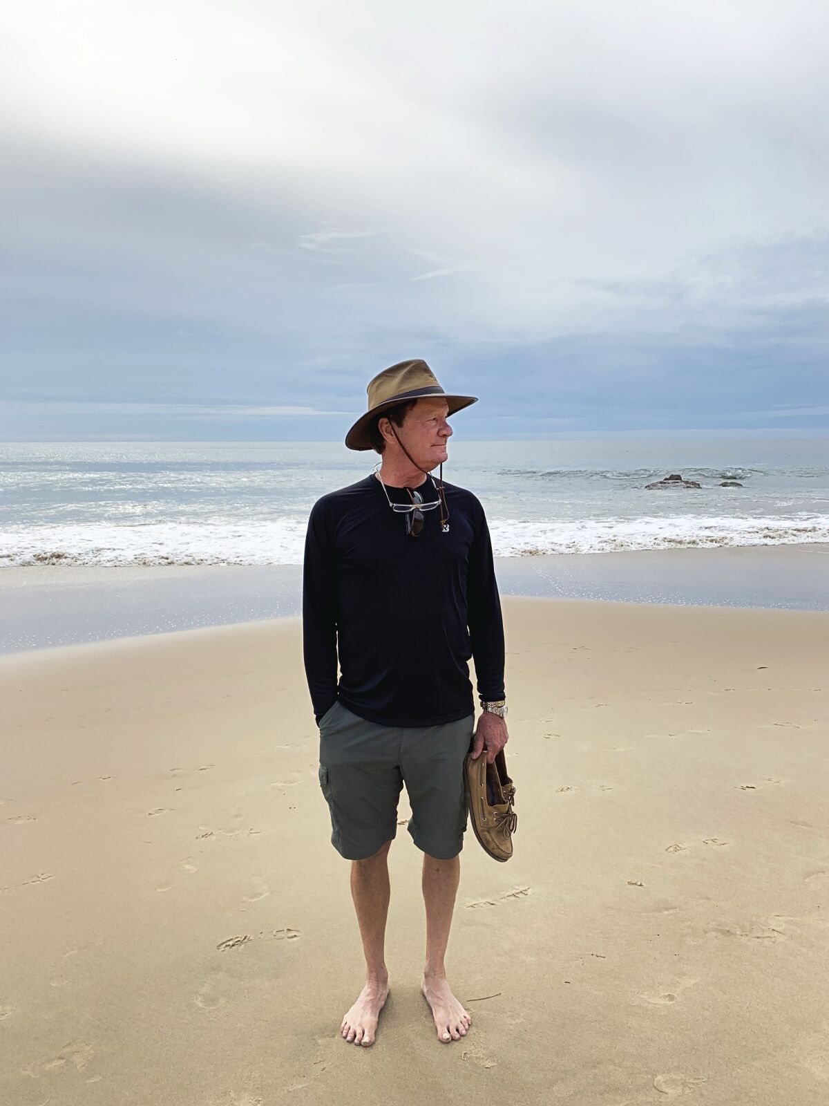 A man in shorts holds his shoes on the beach