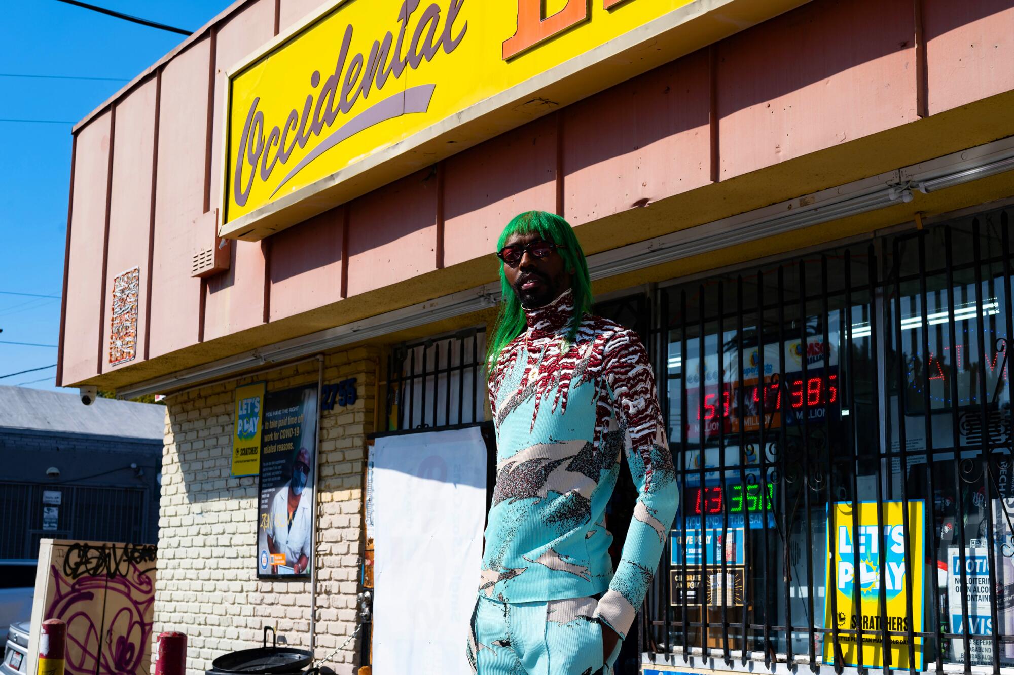 A person in sunglasses and green hair stands in front of a liquor store in a strip mall.