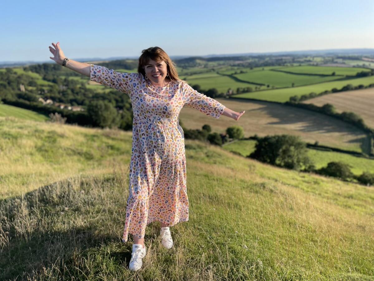 A woman holds her arms out wide atop a hill in the British countryside.