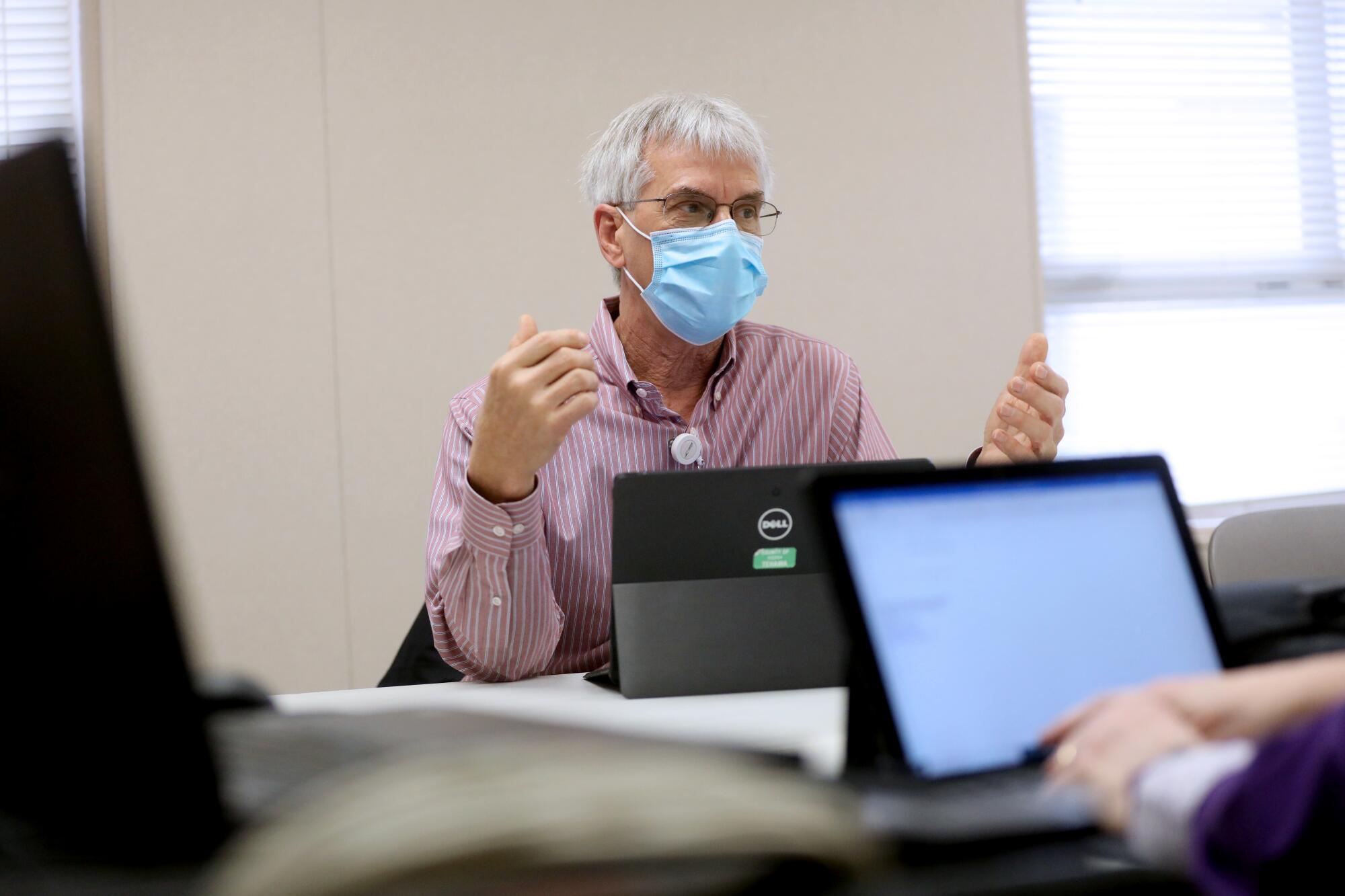 A man in glasses and a blue mask gestures while speaking at a conference table