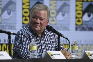 Man, William Shatner, in plaid shirt, sitting in front of mic on panel at comic con, looking to the left, smiling 