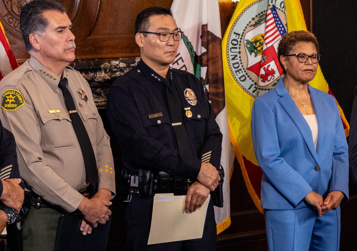Two men in law enforcement uniforms with badges stand with a woman in a pants suit.