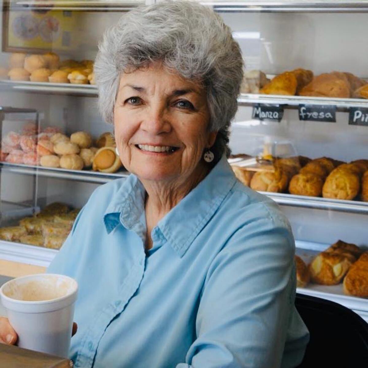 A smiling woman holds a cup near a display of baked goods