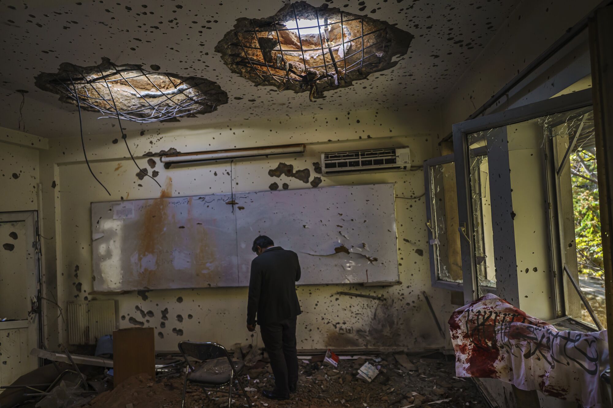 A man stands, hanging his head, inside a classroom with broken windows, holes in the ceiling and debris on the floor.