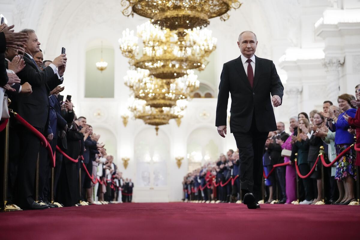 Vladimir Putin walks down a red carpet as people watch behind velvet ropes.