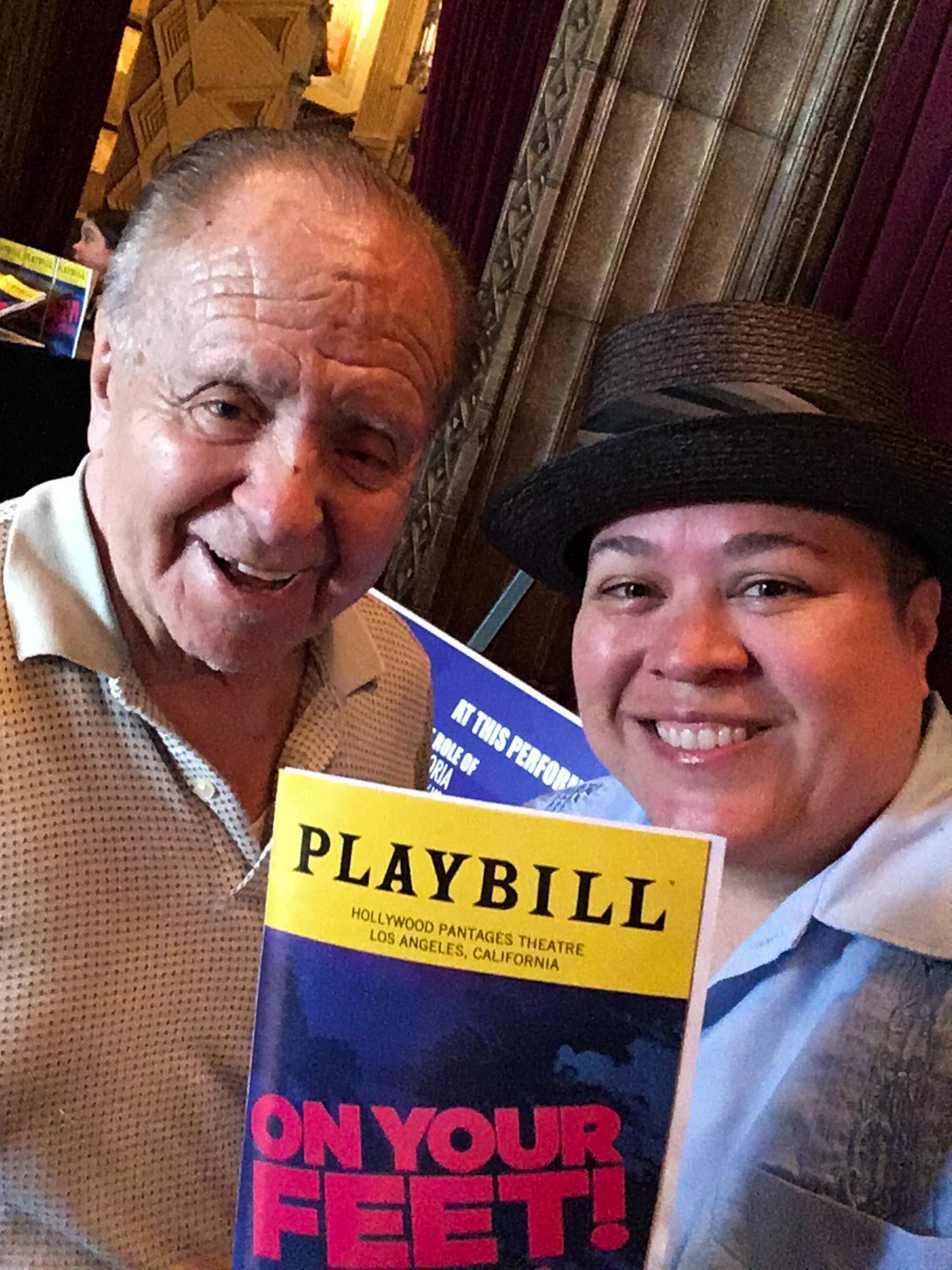 An older man, left, and younger person smiling as they hold up a Playbill for "On Your Feet!"