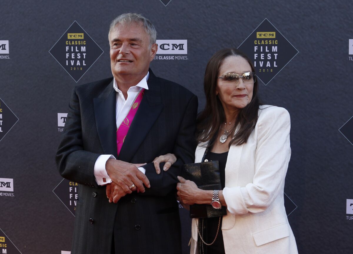 Leonard Whiting, in a black suit, links arms with Olivia Hussey, in a white suit, as the two pose with slight smiles.