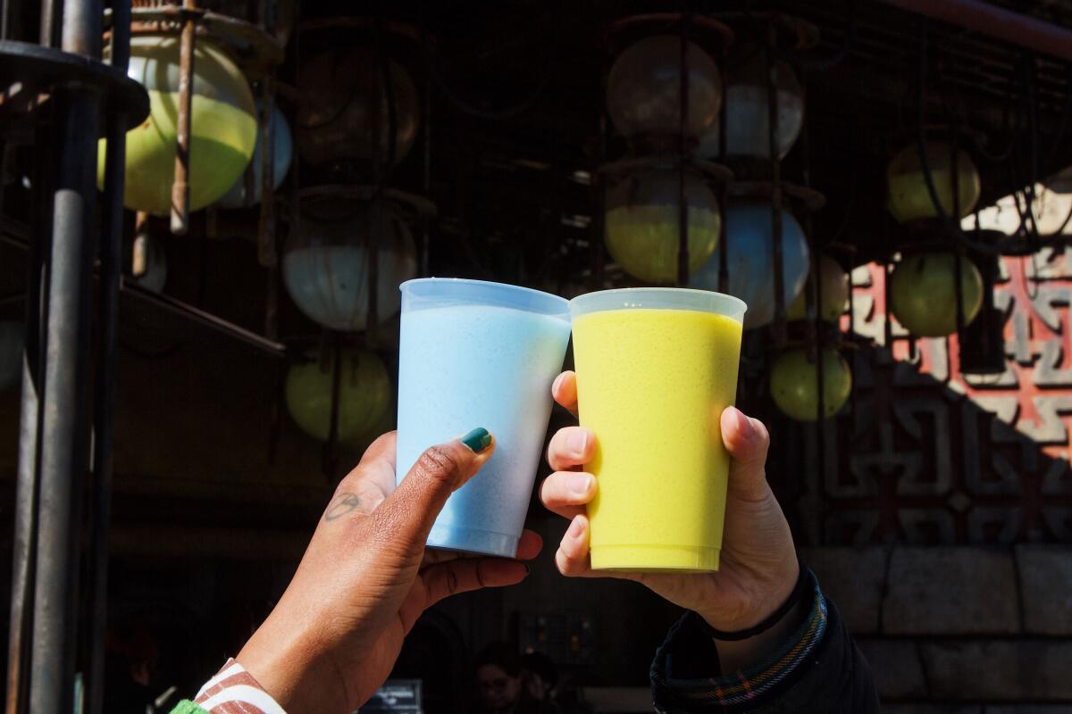 Two hands hold up blue milk and green milk to cheers in front of the Milk Stand.