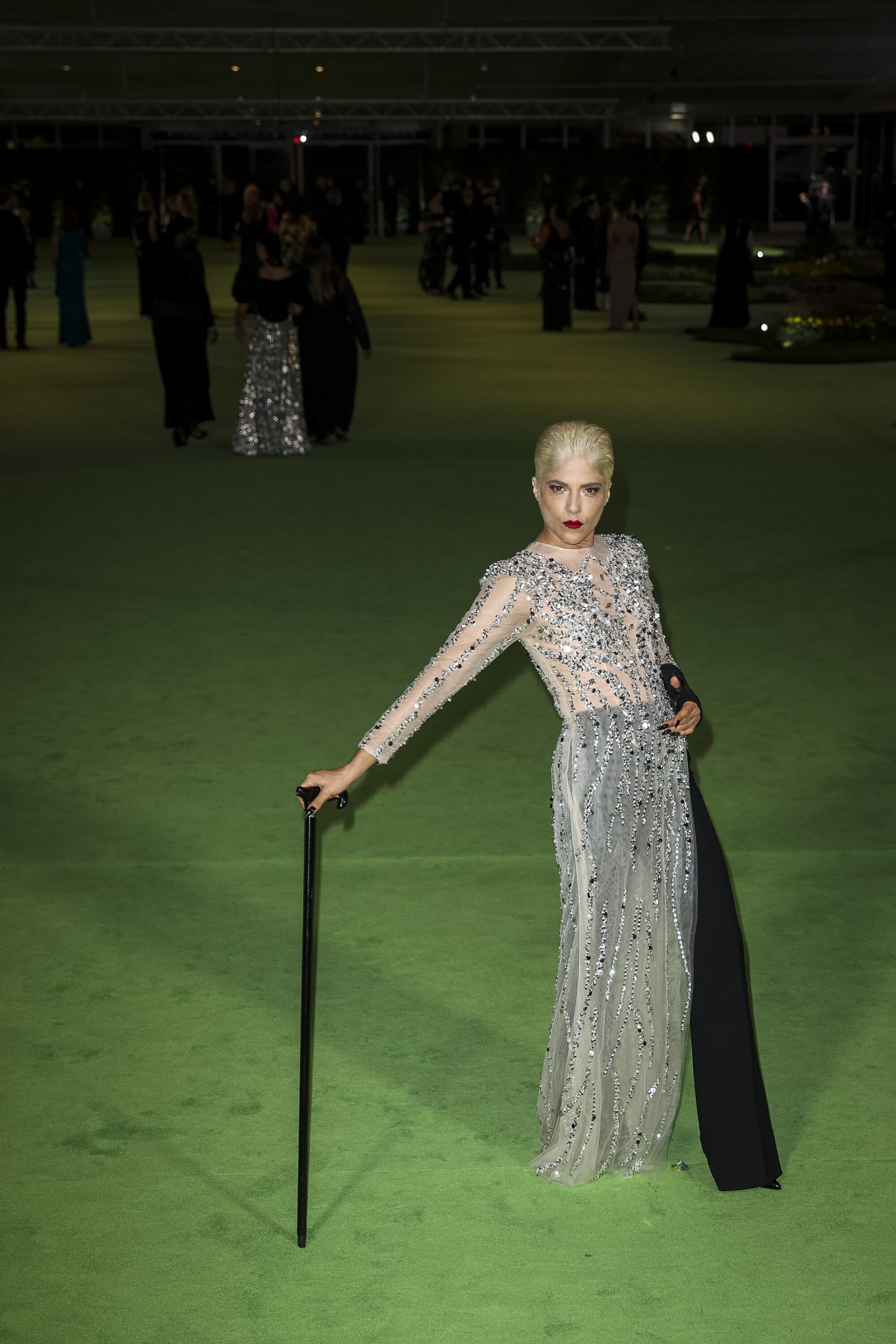 A woman in a silver dress and black pants posing with a cane on a green carpet