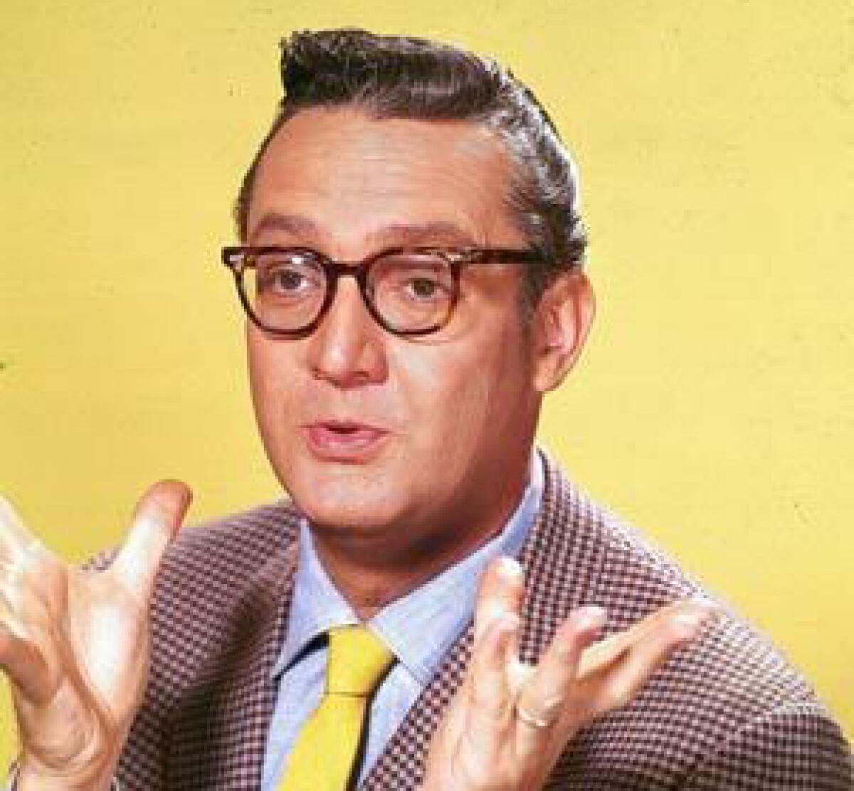 A man with short, dark hair, horn-rim glasses and a checked suit gestures while speaking.