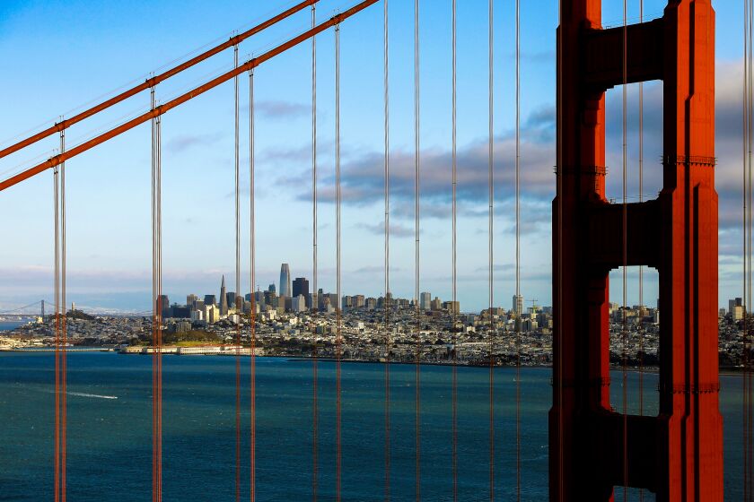 The skyline of downtown San Francisco is visible through the cables that support the Golden Gate Bridge on a sunny day
