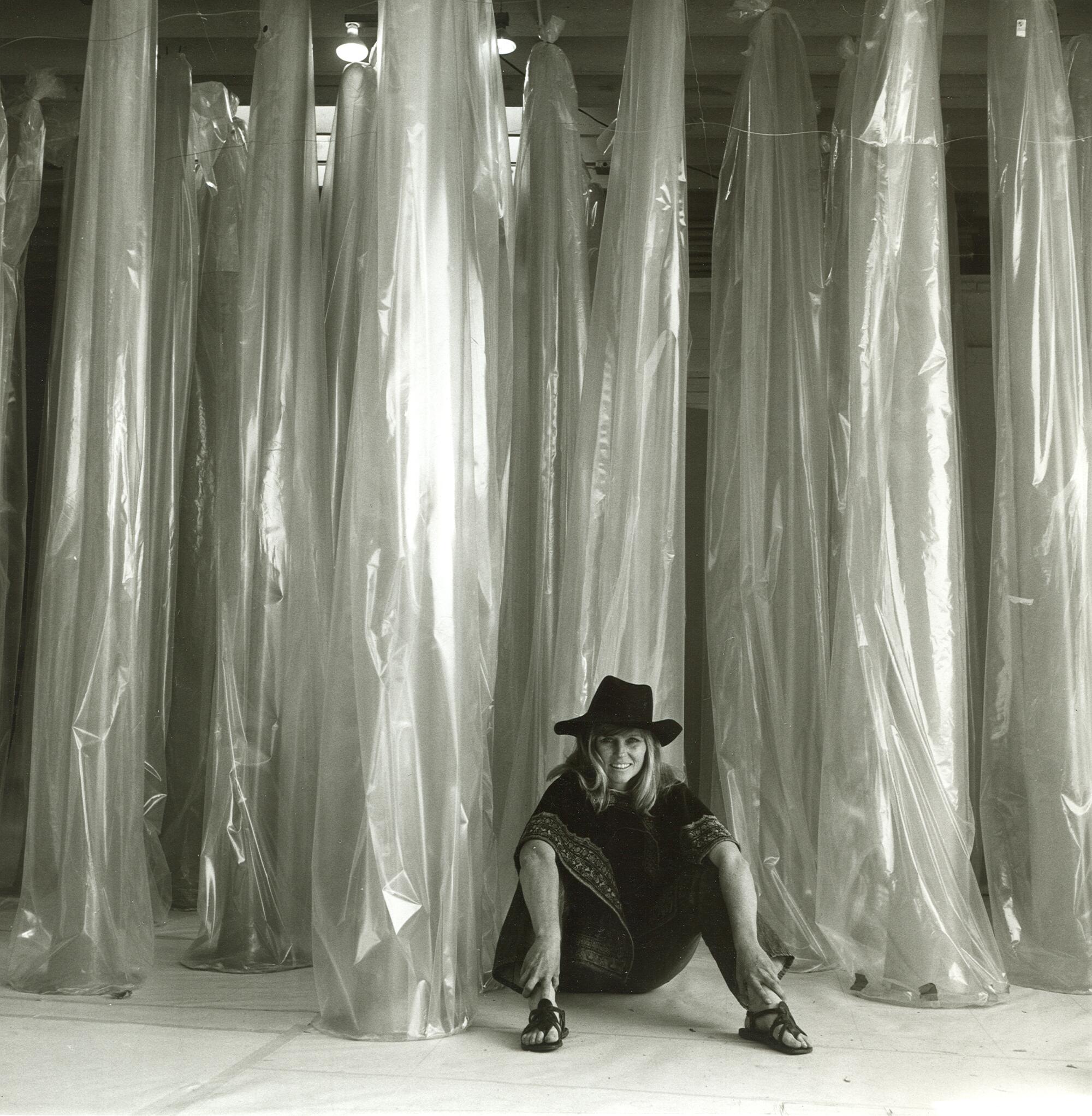 A vintage photo shows Barbara T. Smith wearing a '70s poncho and hat, sitting by a towering installation of plastic tubes 
