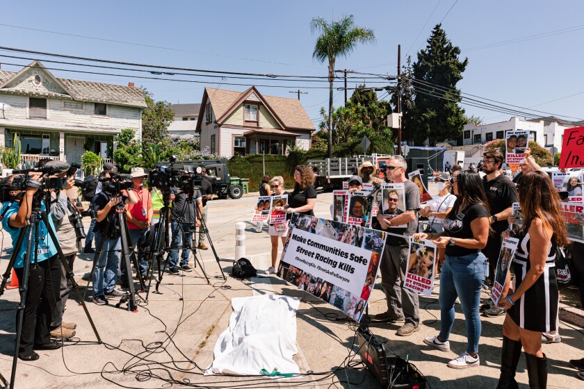 TV cameras pointed at a crowd of people with protest signs and banners, standing behind a white sheet covering a fake body