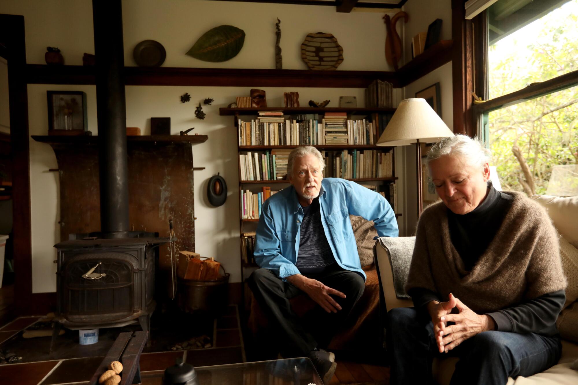 A man and woman in their 70s sit in an eclectic home filled with art and books.