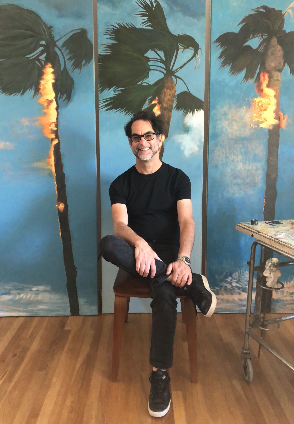 Artist Perry Vásquez sitting in front of his painting of burning palm trees