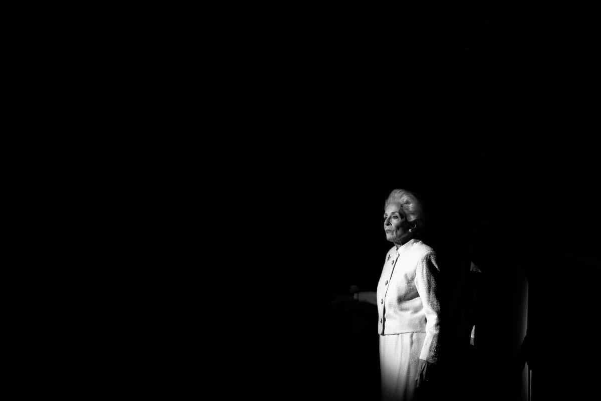 A woman wears a white jacket and skirt on a darkened stage.