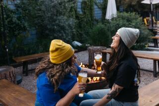 Two people laugh and drink beer around a fire pit.