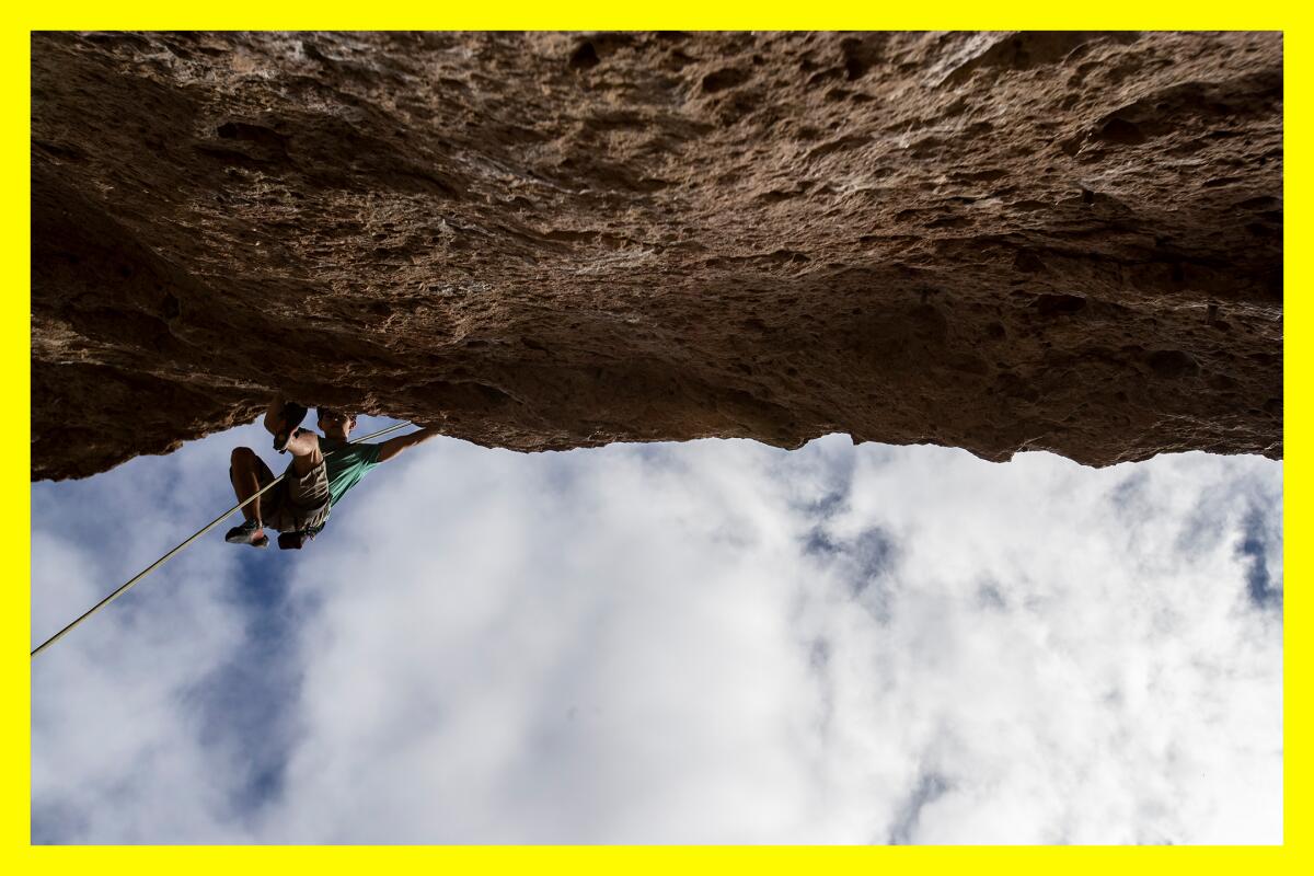 A climber hangs from a rocky wall. Cloudy blue skies are seen above them.