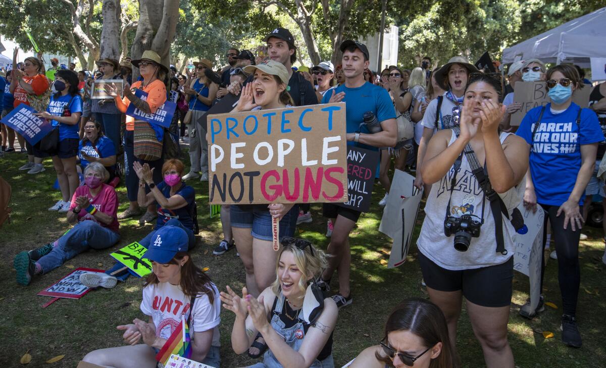 A crowd of demonstrators standing and sitting, one holding a handmade sign reading, "Protect people, not guns."