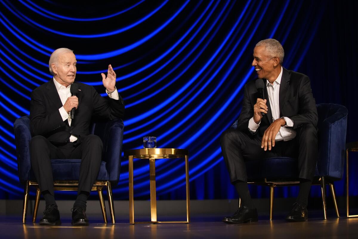 President Biden gestures while speaking with a smiling former President Obama as they sit in front of vivid blue draping.