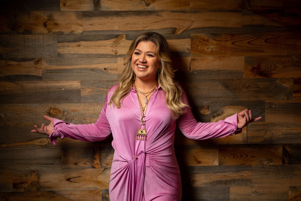 Kelly Clarkson wears a pink dress and stands with her arms out against a brown wall.