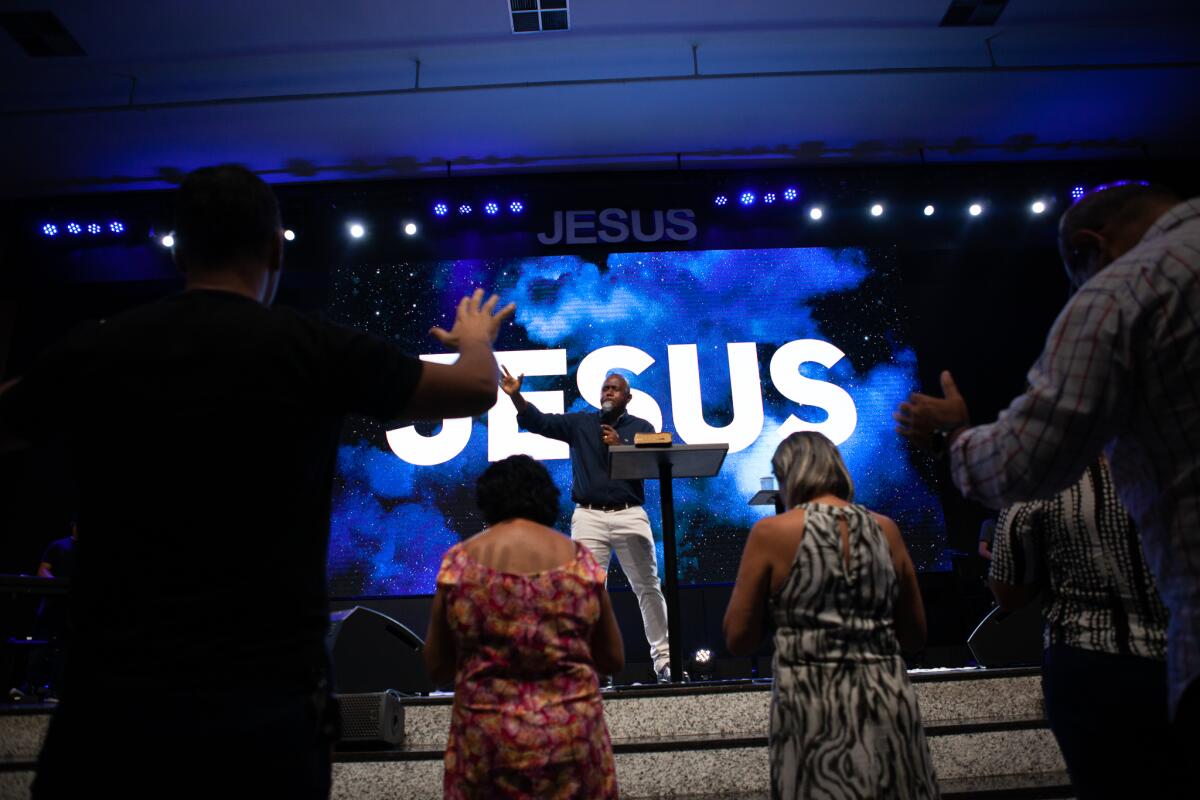 A man standing onstage with the word Jesus behind him speaks to people standing before him 