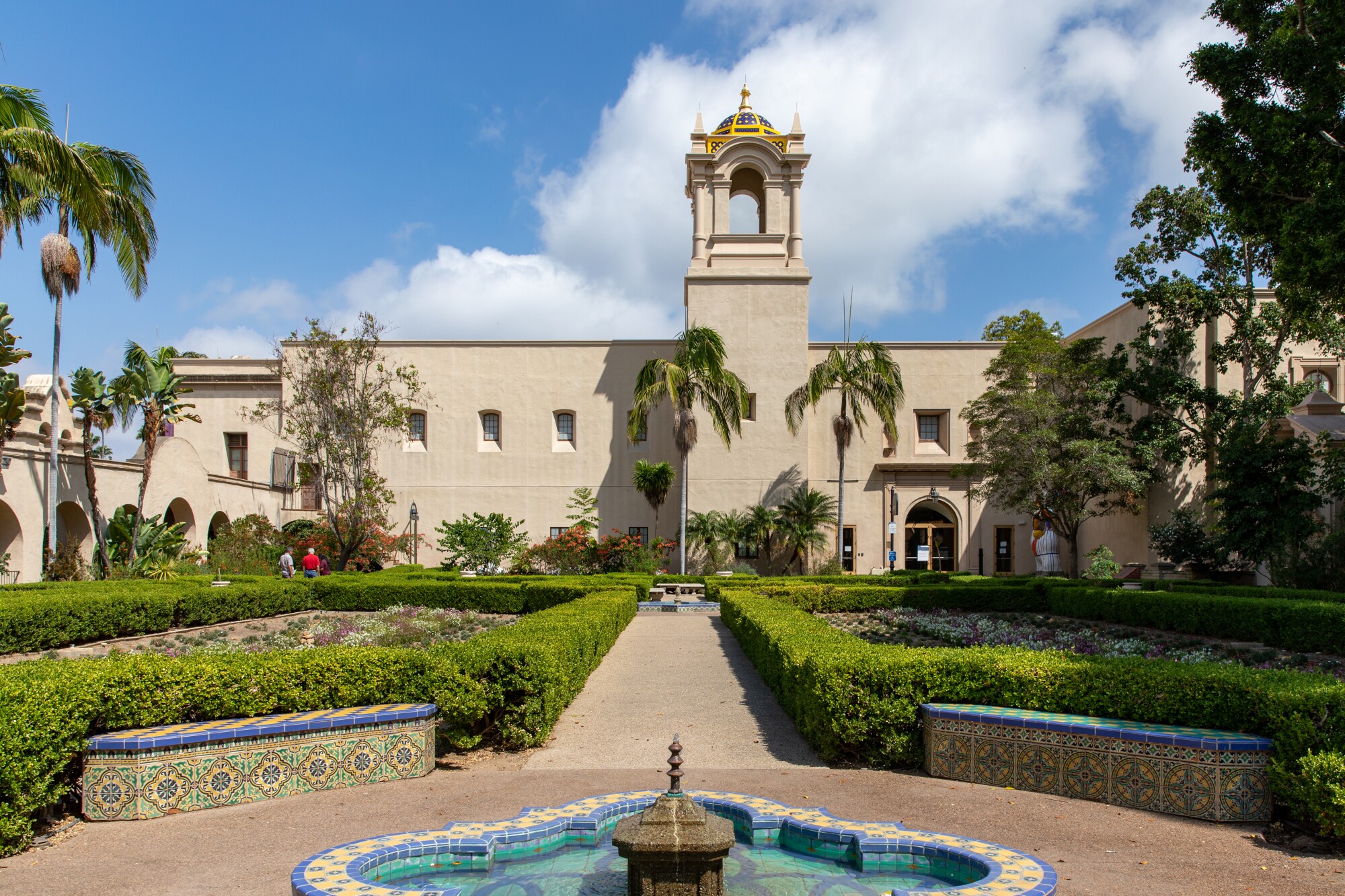 A fountain and a European-style garden can be seen before a multi-story Spanish-style building with a bell tower