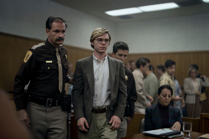 Evan Peters dressed as Jeffrey Dahmer being escorted out of a courtroom by a police officer