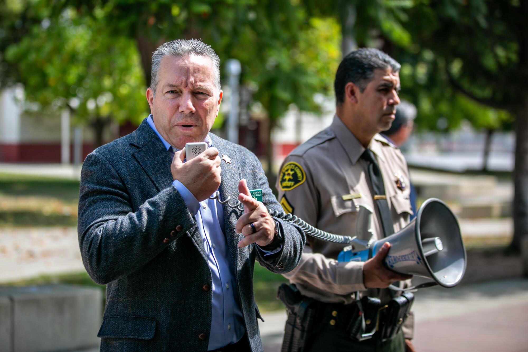 Sheriff Alex Villanueva, left, holding a microphone as another deputy holds a speaker