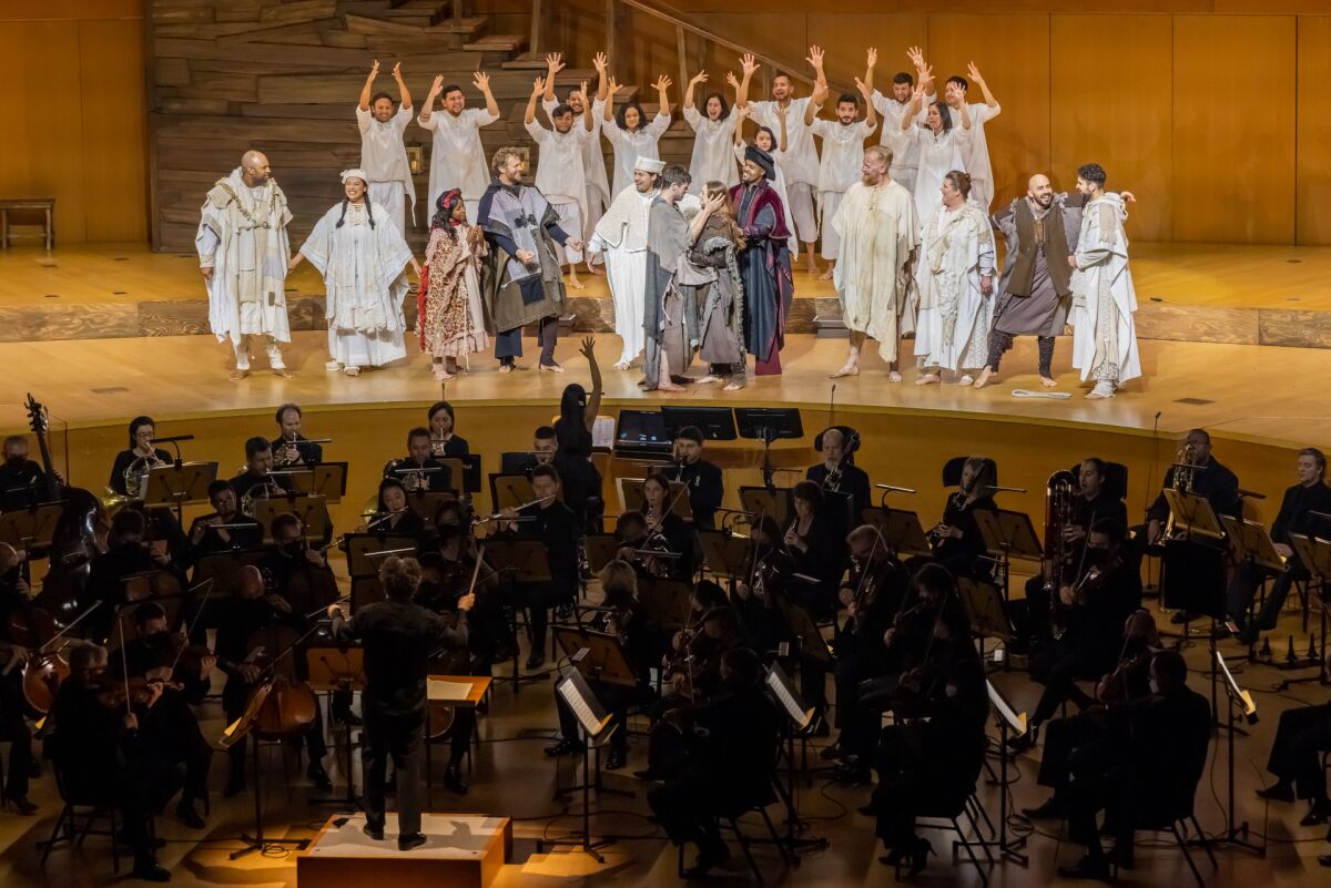 An orchestra performs as people in costume on stage move and lift up their arms.