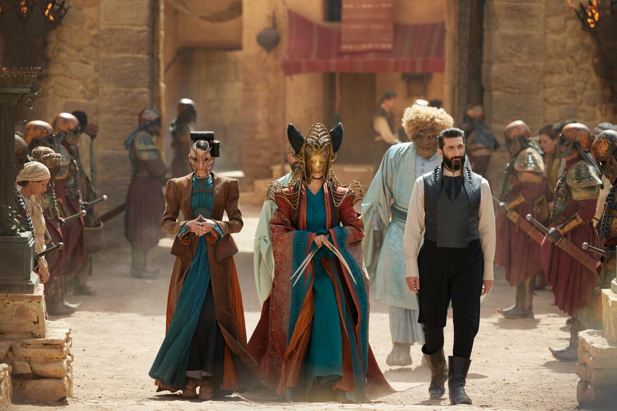 Two masked women and a bearded man walk among soldiers and onlookers on a dusty street.