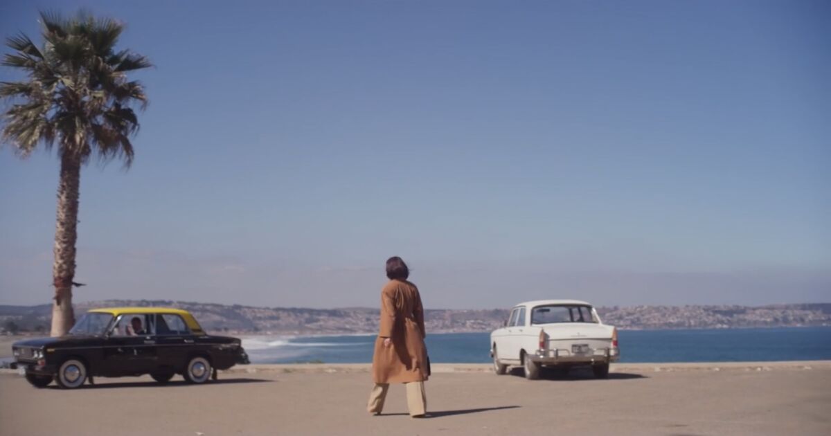 a woman in a parking lot with two old cars looking out on the ocean