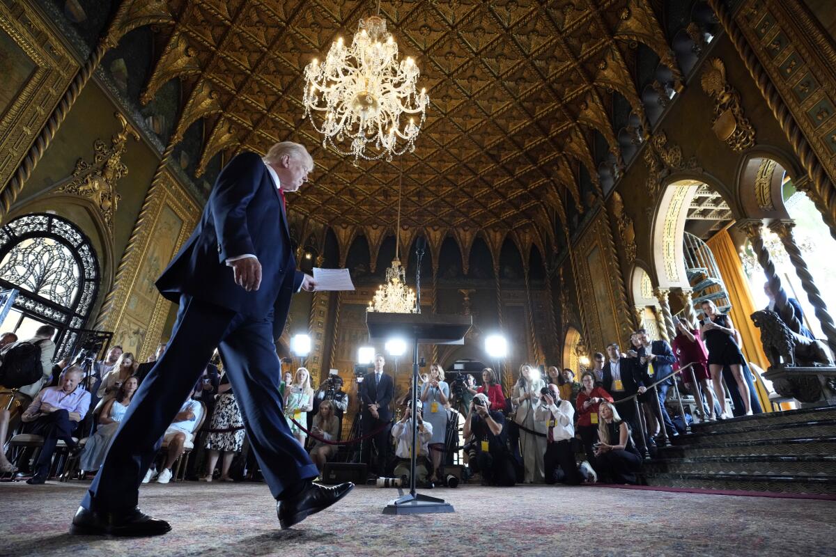 A man in a dark suit walks into a room with people gathered beneath a chandelier