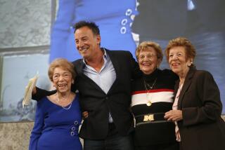 Bruce Springsteen in a dark suit jacket and a suit shirt surrounded by three older women in various formal clothing