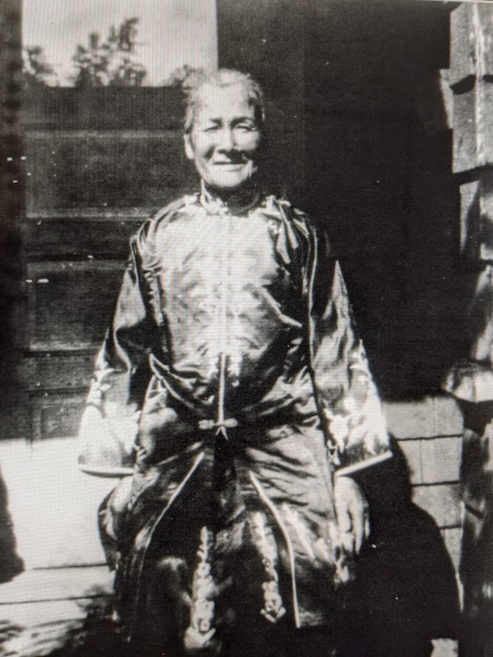 A historic black-and-white photo of an older woman in traditional Chinese dress standing on her porch