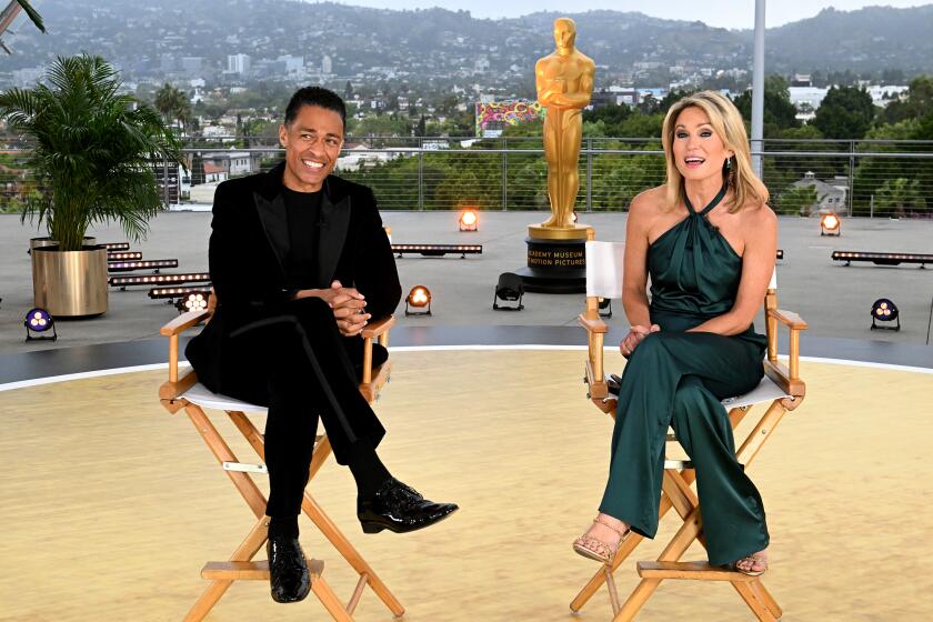 T.J. Holmes in a black outfit sitting next to Amy Robach in a sleeveless jumpsuit sitting in chairs at the Academy Museum