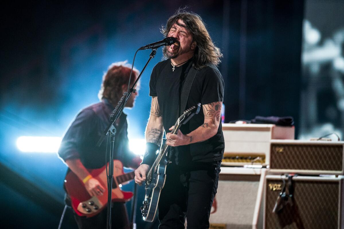 A man with long hair sings into a microphone and plays guitar onstage