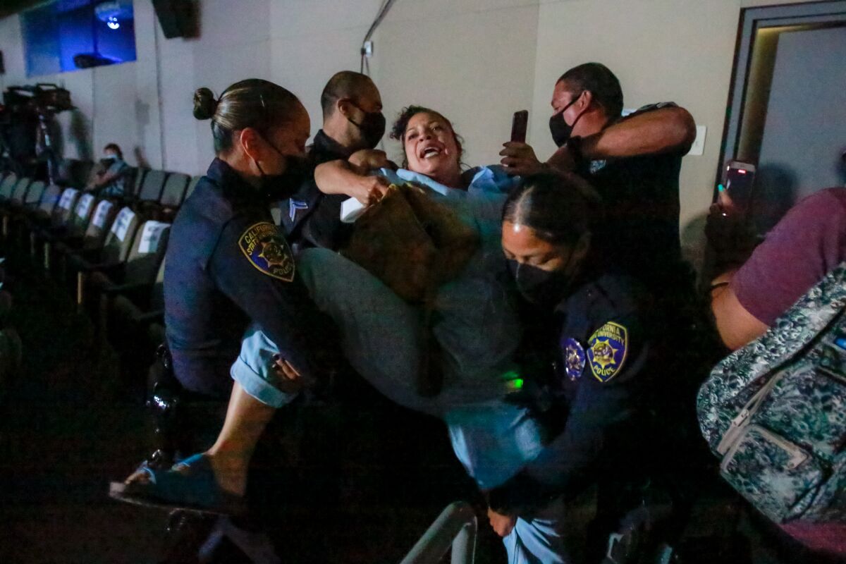 A woman struggles as she is held up and carried, by both arms and legs, by police officers in masks.