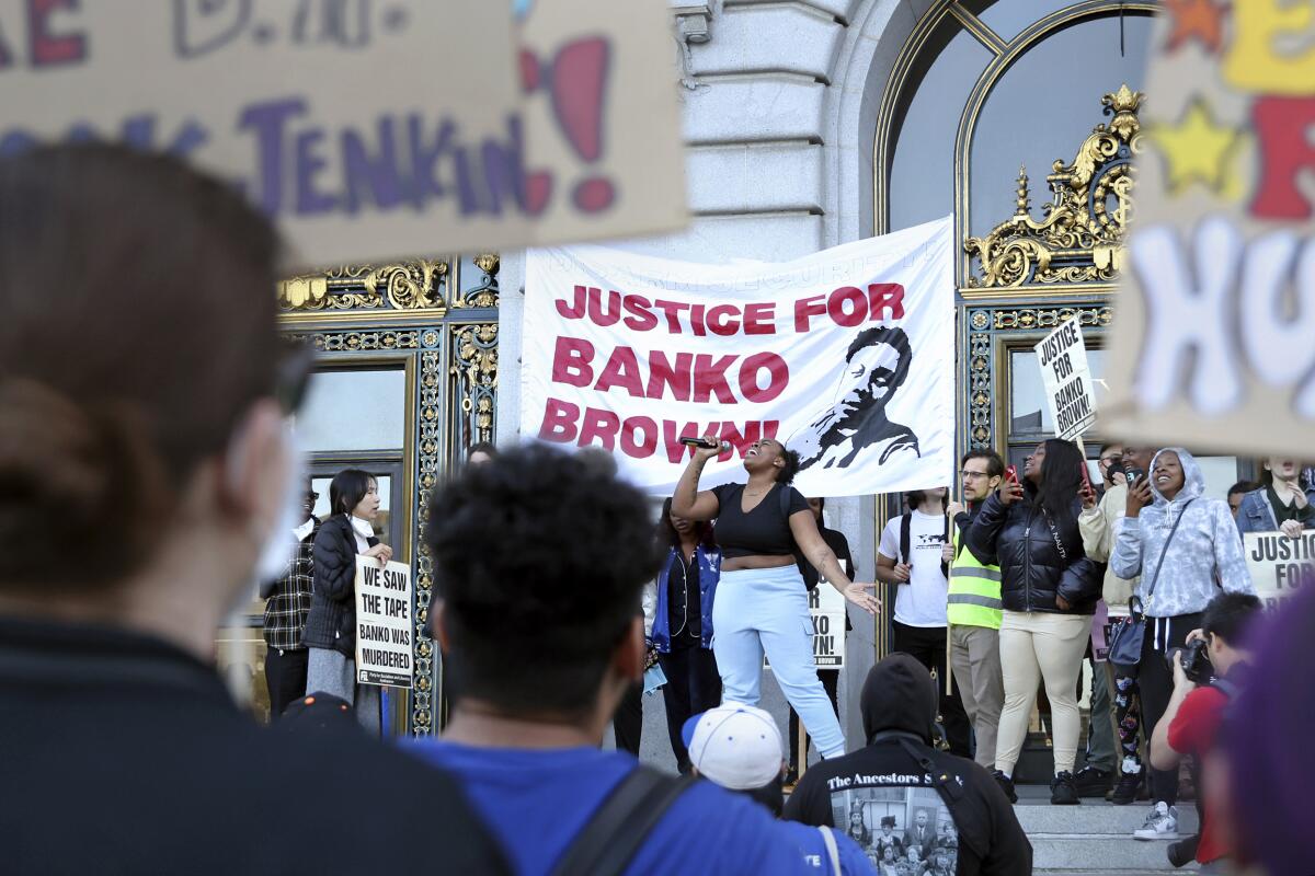 People, some holding homemade signs, gather around a woman speaking in front of a banner reading "Justice for Banko Brown”