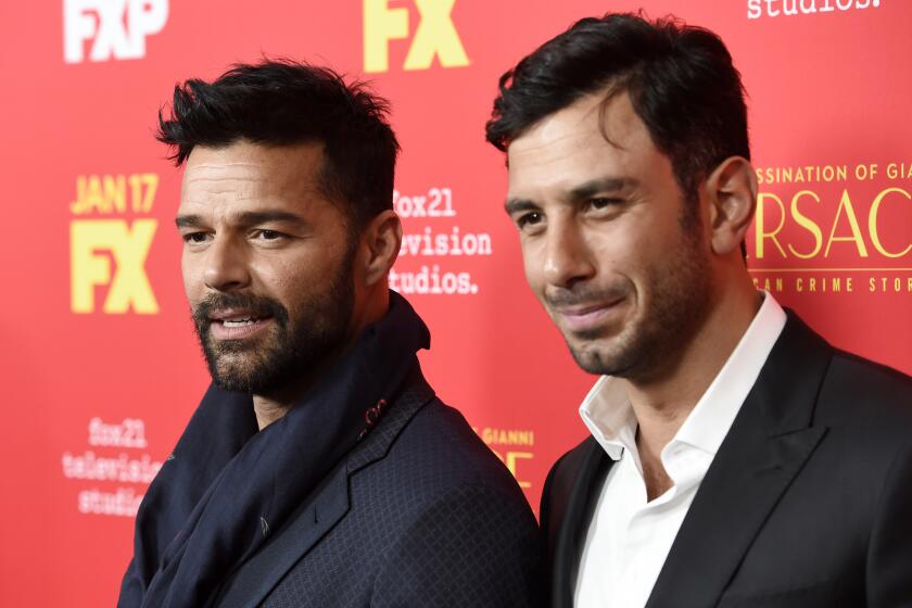 Ricky Martin in a black suit and scarf standing next to husband Jwan Josef in a suit against a red backdrop