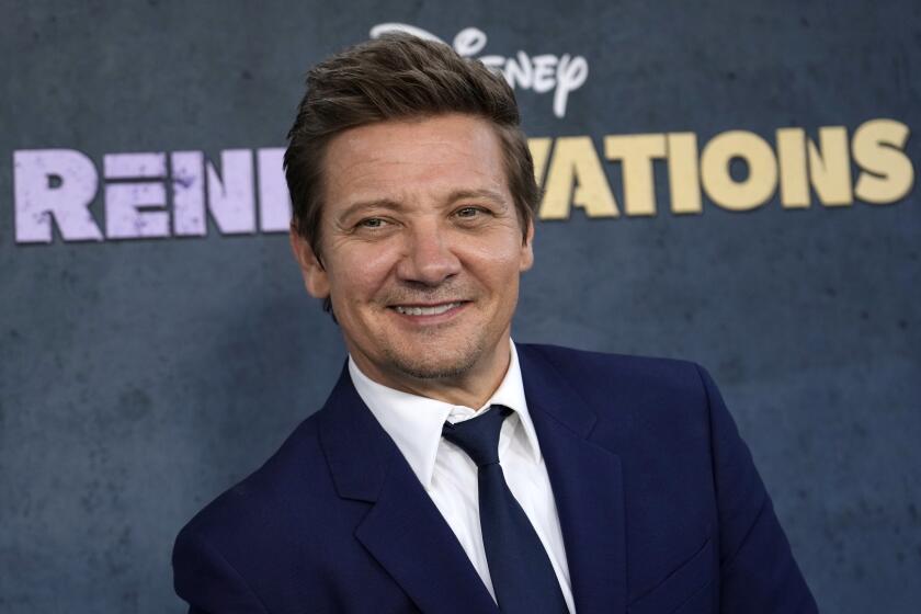 Jeremy Renner in a blue suit and tie smiling and looking to the side