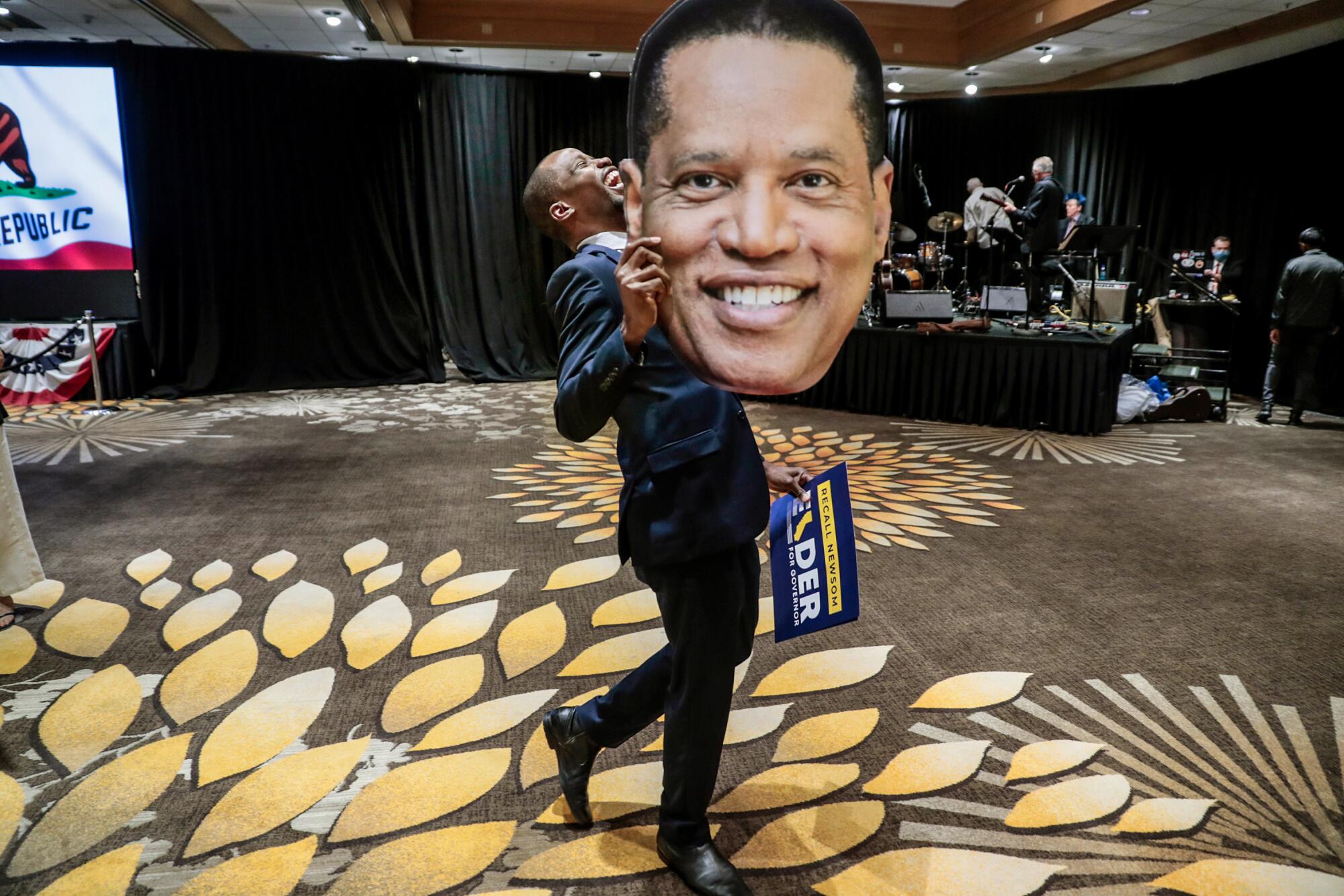 A man in a suit laughs as he displays a large cutout depicting a man's face 