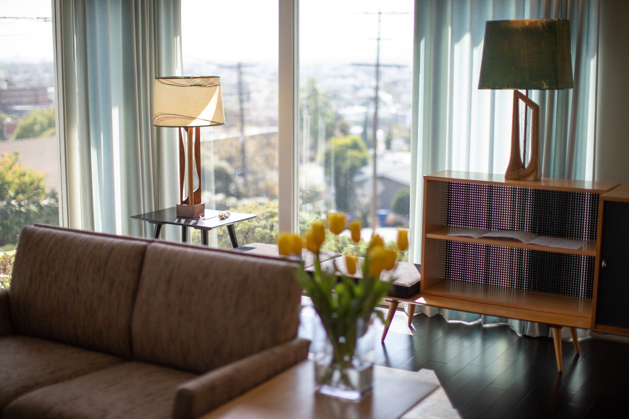 A Modernist living room has two lamps resting, respectively, on a table and a wooden cabinet as sun streams in a window