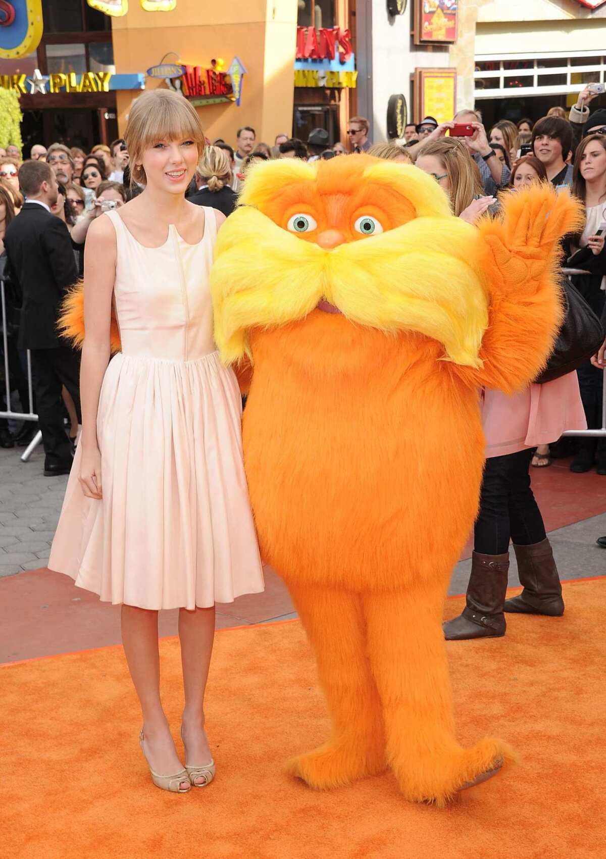 Taylor Swift stands in a pink dress with a fuzzy orange character.